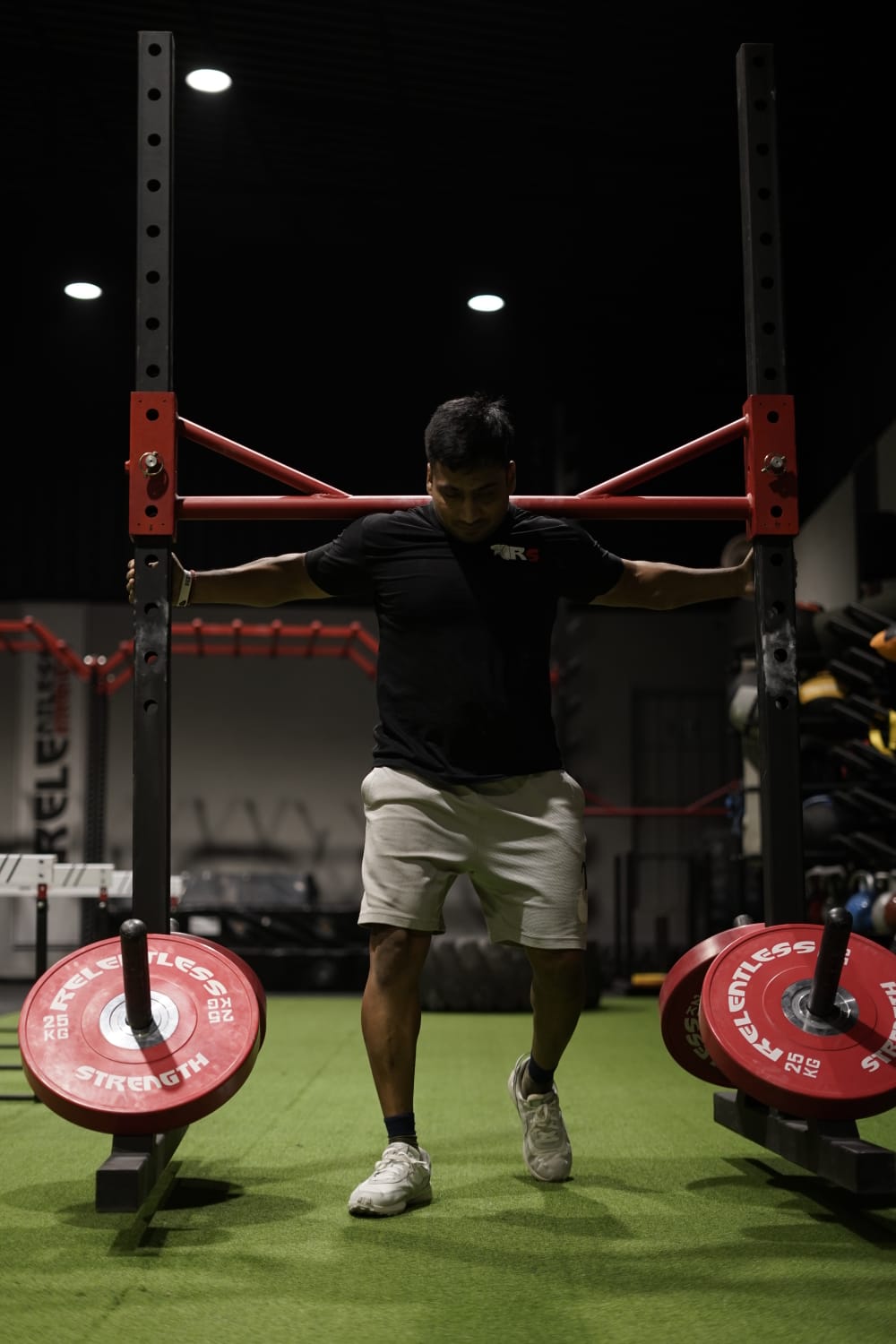 Man performing weighted sledgehammer carry exercise using a metal frame with red weights in a gym with artificial turf.